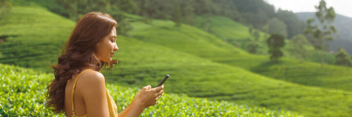 Traveler woman using smartphone during her travel on famous nature landmark. Profile portrait of charming young adult girl with mobile phone against the tea plantations in Nuwara Eliya, Sri Lanka