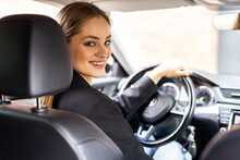 Young beautiful brunette woman sits on drivers seat in a modern car, holding steering wheel turned around and looking into the camera with smile. Travel concept