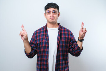 Wall Mural - Portrait of a young handsome Asian man wearing casual shirt and glasses doing fingers crossed, wishing and praying for miracle.