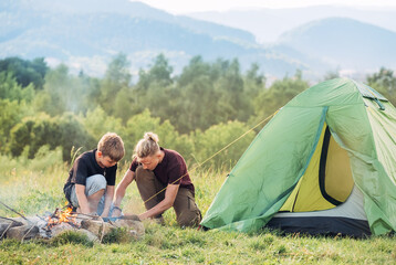 two brothers teens setting up the green tent next to campfire. outdoor active time spending or campi