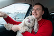 Happy man hugs his white lapdog, smiles and points his hand forward. Owner of fluffy Chinese crested dog travels by car. Travel with pet concept.