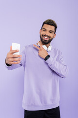 Wall Mural - happy african american man with wireless headphones taking selfie while showing peace sign isolated on purple.