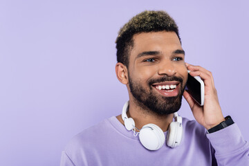 Wall Mural - happy african american man in wireless headphones talking on smartphone isolated on purple.