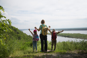 mom and two kids with backpack walk near the river. Staycations, hyper-local travel, family outing, getaway, natural environ. Concept of friendly family. Family spends summer time together on a hike