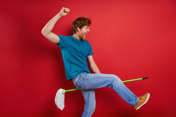 Playful young redhead man using broom like guitar while against red background