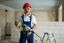 Professional Construction Worker In Uniform Standing With Rotary Hammer Drill. Portrait Of Contractor In Hardhat And Overalls Posing With Jackhammer Near Step Ladder And Masonry Indoors.