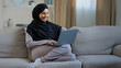 © Yuliia - Friendly arabian muslim woman girl in hijab sit on cozy sofa talk video call with friend greeting at computer webcam take interview distance virtual communication in social networks use wi-fi at home