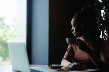 Daydreaming african afro american young businesswoman, manager drinking coffee in cafe, looking out window and working on laptop. Multicultural female on remote freelance occupation in creative office