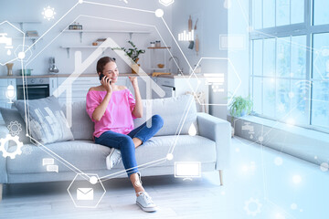 Wall Mural - Happy young woman talking by mobile phone on sofa at home. Modern technology and smart home automation