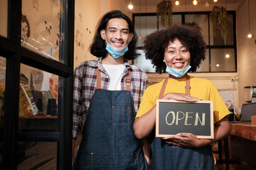 Wall Mural - Two young startup barista partners stand at casual cafe door, write and show open sign before taking off face masks, happy and cheerful smiles with coffee shop service jobs after COVID-19 lifestyle.