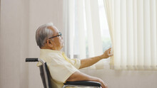 Old Asian Man Sits In A Wheelchair And Looks Out The Window. Health Care And Insurance, Retirement Concept