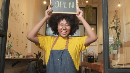 Wall Mural - One African American female startup barista stands at casual cafe door, looks at camera, and shows open sign, happy and cheerful smiles with coffee shop service jobs, and new business entrepreneurs.