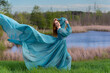 One red haired woman wearing long flowing blue gown in green meadow with a lake.  Magical, fairytale concept.