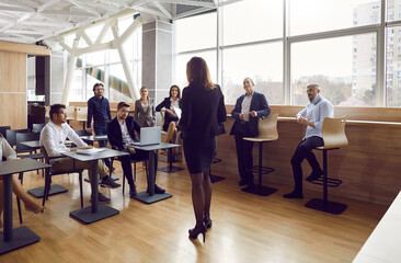 Wall Mural - Group of diverse business people listen to speech by female speaker at business meeting. Experienced business lady speaks in front of audience in loft office. Concept of business speeches.