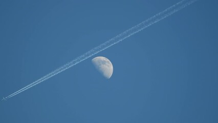 plane flying high in clear sky passing close by half moon in background, real time footage