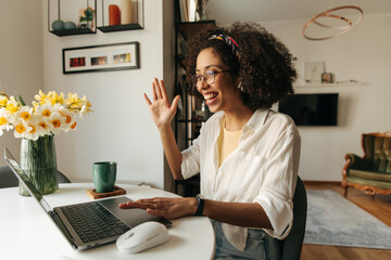 Friendly young african girl smiles and waves at laptop camera sitting at table. Brunette with afro hair wears white shirt. Concept of use