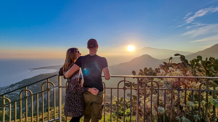 Wall Mural - Hugging couple watching beautiful sunset behind volcano Mount Etna near Castelmola, Taormina, Sicily, Italy, Europe, EU. Clouds with vibrant red orange colors. Silhouette of people during twilight