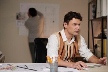 Canvas Print - Concentrated architect working on computer when his colleague putting blueprint on wall in backg