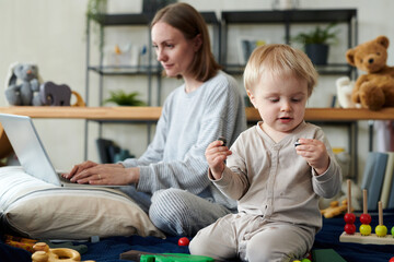 Cute little child playing toys on bed with his working mother using laptop for online work in background