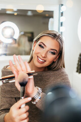 Wall Mural - Happy young woman streaming a beauty makeup vlog from home or workshop. Beautiful online content creator cosmetician applying makeup and explaining some work tools. Vlogging and online channel work.