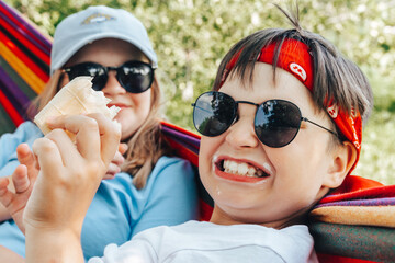 Cheerful teenagers in stylish glasses eating sweet ice cream in hammock at summer vacation.Happy Children licking icecream refreshing in heat.Cool off, Summer food.