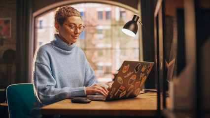 Fototapete - Young Attractive Woman Working from Home on Laptop Computer with Stickers in Sunny Stylish Loft Apartment. Creative Designer Wearing Cozy Blue Sweater and Glasses. Urban City View from Big Window.