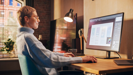Fototapete - Young Attractive Woman Working from Home on Desktop Computer in Sunny Stylish Loft Apartment. Creative Designer Wearing Cozy Blue Sweater and Glasses. Urban City View from Big Window.