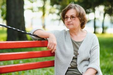 Poster - Elegant elderly woman in the shirt is sitting on the bench in a park on a warm day