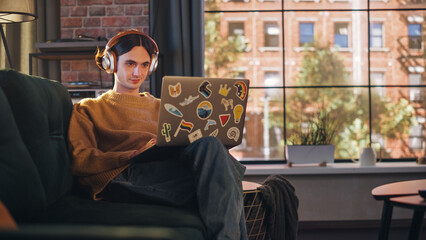 Wall Mural - Young Handsome Man Sitting on Sofa and Using Laptop Computer in Sunny Stylish Loft Apartment. Creative Designer Wearing Cozy Yellow Sweater and Headphones. Urban City View from Big Window.