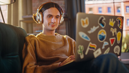 Poster - Young Handsome Man Sitting on Sofa and Using Laptop Computer in Sunny Stylish Loft Apartment. Creative Designer Wearing Cozy Yellow Sweater and Headphones. Urban City View from Big Window.