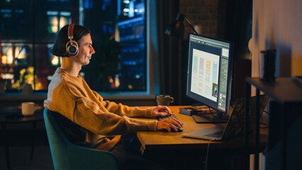 Fototapete - Young Handsome Man Working from Home on Desktop Computer in Stylish Loft Apartment in the Evening. Creative Designer Wearing Cozy Yellow Sweater and Headphones. Urban City View from Big Window.