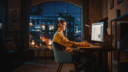 Fototapete - Young Handsome Man Working from Home on Desktop Computer in Stylish Loft Apartment in the Evening. Creative Designer Wearing Cozy Yellow Sweater and Headphones. Urban City View from Big Window.