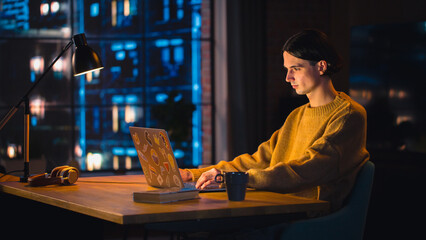 Poster - Young Handsome Man Working from Home on Laptop Computer in Stylish Loft Apartment in the Evening. Creative Person Wearing Cozy Yellow Sweater and Headphones. Urban City View from Big Window.