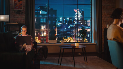 Aufkleber - Young Couple Spending Time at Home, Working on Computers from Their Stylish Loft Apartment in the Evening. Female Resting on the Sofa and Browsing Social Media on Laptop. Man Designing UX Interface.