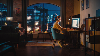 Fototapete - Young Couple Spending Time at Home, Working on Computers from Their Stylish Loft Apartment in the Evening. Female Resting on the Sofa and Browsing Social Media on Laptop. Man Designing UX Interface.