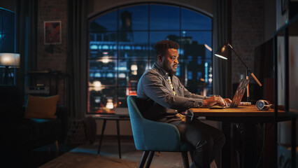 Fototapete - Young Handsome African American Man Working from Home on Laptop Computer in Stylish Loft Apartment at Night. Creative Male Checking Social Media, Browsing Internet. Urban City View from Big Window.