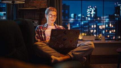 Wall Mural - Young Beautiful Woman Resting on Couch, Using Laptop Computer in Stylish Loft Apartment in the Evening. Creative Female Smiling, Checking Social Media, Stretching. Urban City View from Big Window.