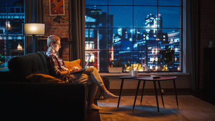 Poster - Young Beautiful Woman Sitting on Couch, Using Laptop Computer in Stylish Loft Apartment in the Evening. Creative Female Smiling, Checking Social Media, Stretching. Urban City View from Big Window.