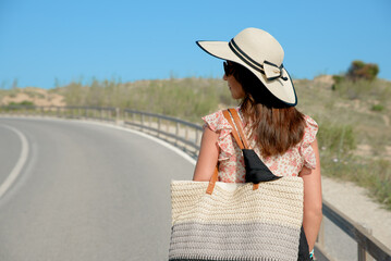 Young woman on vacation walks down a road with a hat and bag