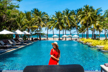 Travel Vacation Woman In Red Dress Enjoying A Summer Vacation Near Swimming Pool In Tropical Resort Near The Beach With Sea View. Carefree Girl Tourist In Hat Relax In Luxury Resort, Thailand