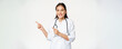 Smiling female asian doctor in medical uniform, pointing fingers and looking left at advertisement, copy space promo, standing in robe against white background