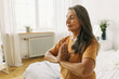 © shurkin_son - Woman of 60s doing namaste gesture with palms pressed against each other, keeping eyes closed dressed in yellow sport t-shirt, having long hair, sitting on bed against window, reaching harmony