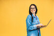 Leinwandbild Motiv Portrait of happy young beautiful surprised woman with glasses standing with laptop isolated on yellow background. Space for text.