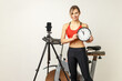 Time for sport. Sporty woman with perfect shape standing near exercise and tripod with phone, holding wall clock, wearing sports tights and red top. Indoor studio shot on gray wall background.