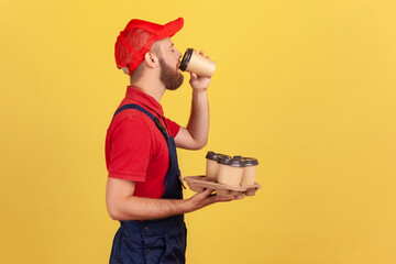 Wall Mural - Side view portrait of bearded handy man standing with disposable cups and drinking hot beverage, needs energy for continue working. Indoor studio shot isolated on yellow background.