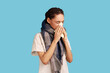 Unhealthy woman feels unwell, blows nose in tissue, suffers from running nose, cold symptoms or allergy, sneezes very often, wearing white shirt. Indoor studio shot isolated on blue background.