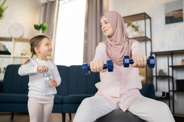 Wall Mural - Focus on dumbbells, young Arab family doing sports in a bright living room at the weekend. Happy free time, healthy lifestyle concept.