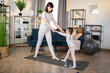 © sofiko14 - Full length portrait of woman wearing white t shirt and leggins doing sport exercises at home with her daughter, standing on mat, stretching body, training together.