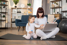Mum And Daughter Staring On Laptop Screen, Holding Bottles Of Water. Have A Break. Relaxing Together. Fitness At Home. Holiday Leisure. Body Balance. Laying On A Gym Carpet. Family Relationship.