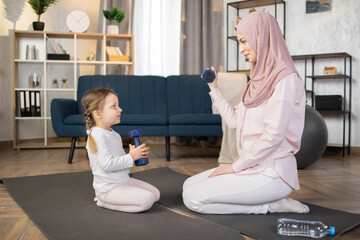 Wall Mural - Cute little girl doing sports with her Muslim mother in a hijab sitting on a mat and doing exercises with dumbbells in living room at home.